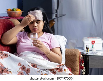An Indian Asian woman suffering from fever drinking water and applying wet cloth on forehead while lying on sofa