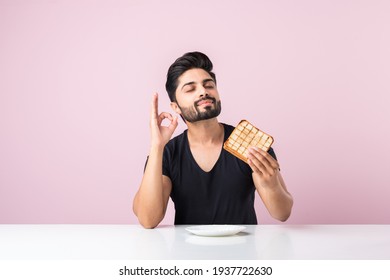 Indian Asian young bearded man eats bread sandwich while sitting in kitchen or dining table. Showing or presenting