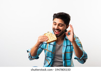 Indian Asian young bearded man eats bread sandwich while sitting in kitchen or dining table. Showing or presenting
