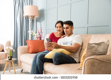 Indian asian young couple using smartphone together while sitting on sofa or couch and modern interior space or home