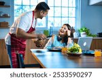 An Indian Asian young husband in an apron lovingly presents a homemade noodles dish to his wife, who is busy working from home at the dining table with her laptop