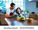 An Indian Asian young husband in an apron lovingly presents a homemade noodles dish to his wife, who is busy working from home at the dining table with her laptop