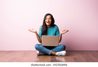 Indian Asian Young Woman Or Girl Sitting With Laptop On Her Lap Against Pink Wall On Wooden Floor