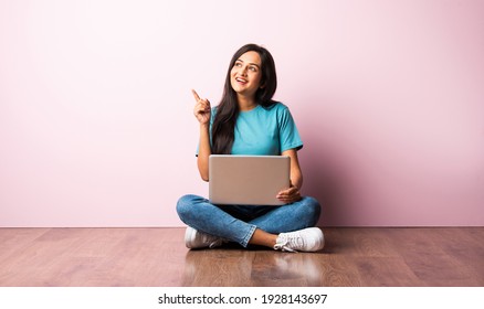 Indian Asian Young Woman Or Girl Sitting With Laptop On Her Lap Against Pink Wall On Wooden Floor