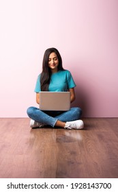 Indian Asian Young Woman Or Girl Sitting With Laptop On Her Lap Against Pink Wall On Wooden Floor