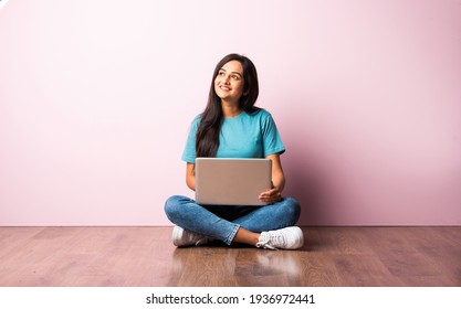 Indian asian young woman or girl sitting with laptop on her lap against pink wall on wooden floor