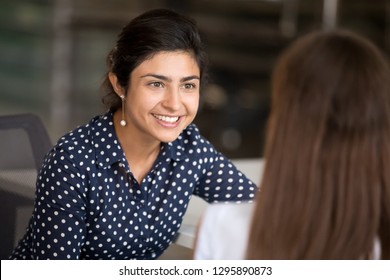 Indian Attractive Positive Woman Talking With Colleague Sitting At Desk In Office Room, Focus On Hindu Female. Diverse Millennial Smiling Employees Have Break Communicating Together At Coworking Area