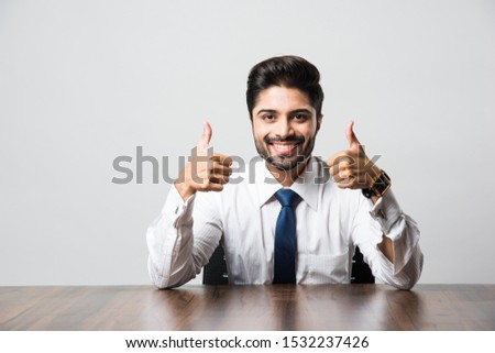 Indian Bearded Male businessman celebrating success with thumbs up or raising fist while sitting at table / desk in office