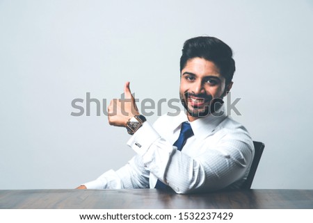Indian Bearded Male businessman celebrating success with thumbs up or raising fist while sitting at table / desk in office