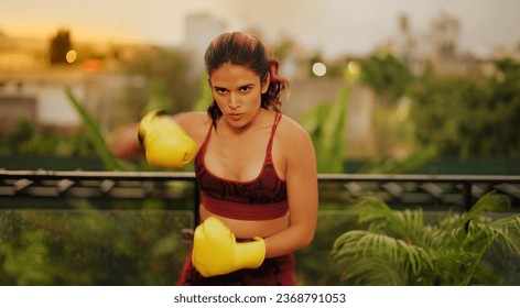 Indian beautiful angry young woman in sportswear doing sport training punching on air at outdoor home. Healthy athletic boxer female standing in boxing gloves learn exercise beating workout at house - Powered by Shutterstock