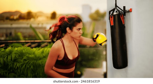 Indian beautiful angry young woman in sportswear doing sport training punching on bag outdoor home. Healthy athletic boxer female standing in boxing gloves learn exercise beating workout at house - Powered by Shutterstock