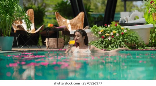 Indian beautiful woman standing in swimming pool edge hold glass drinking juice at outdoor home. Happy female bathing in luxury resort hotel doing relaxation pose in blue water enjoy summer vacation