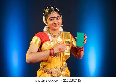 Indian bharatanatyam dancer showing green screen mobile phone by pointing finger while looking at camera - concept of app advertisment, promotion and artist.