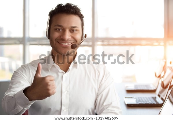 Indian black call center operator\
shows thumb up. Portrait of smiling cheerful man in white shirt\
sitting in bright office. Bright windows\
background.