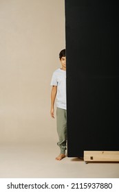 Indian Boy Standing Half Of Him Behind A Black Backdrop Over Beige Background. Standing Straight, Looking At The Camera.