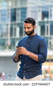 Indian business man, eastern businessman holding cellphone using mobile phone banking app, making e payment, texting message, reading news standing in urban city on modern street outdoors, vertical.