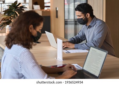 Indian business man student wearing face mask working on laptop safe distancing. Diverse people in facemasks using computers sitting at table with social distance sign in office coworking or cafeteria