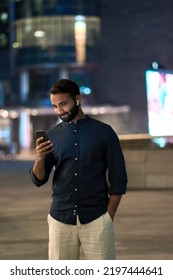 Indian Business Man Wearing Earbuds Standing Outdoors On Night Big City Street Holding Cellphone Using Smartphone Looking At Mobile Phone Watching Streaming Tv Game Or Having Mobile Chat Video Call.