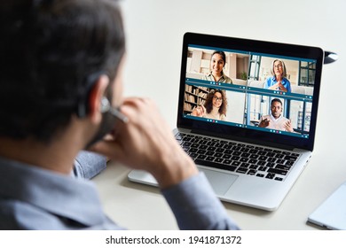 Indian Business Man Wearing Headset Having Virtual Team Meeting On Video Conference Call Using Laptop Work From Home Office Talking To Diverse Group In Remote Online Distance Chat. Over Shoulder View