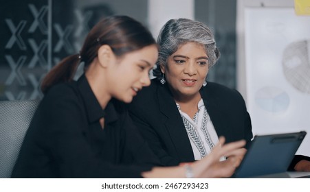 Indian business women sitting on chair using tablet look screen discuss share new project plan ideas job office place. Young adult colleagues old elder female doing work task together indoor workplace - Powered by Shutterstock