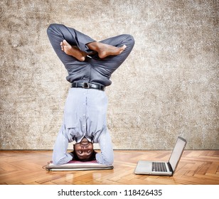 Indian Businessman Doing Yoga Headstand Pose And Looking At His Laptop In The Office At Brown Textured Background