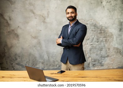 Indian businessman in formal suit stands in modern office near desk with a laptop and looking at camera. Ambitious smiling multiethnic man in smart casual wear with arms crossed on grey background