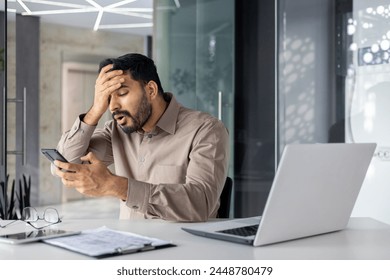 An Indian businessman looking confused and stressed as he examines his smartphone in a modern office setting, with a laptop and documents on the desk. - Powered by Shutterstock