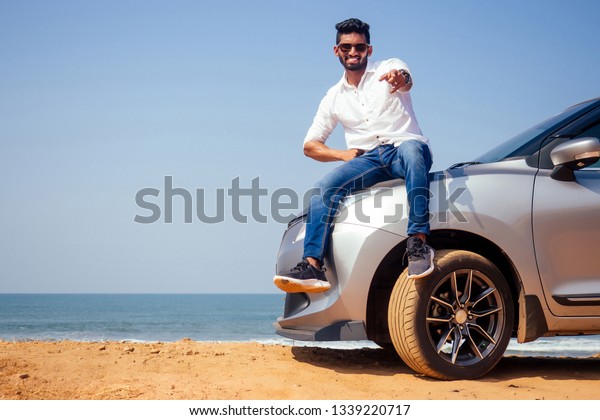 Indian\
businessman standing near car outdoors on sea beach summer good\
day.a man in a white shirt and snow-white smile rejoicing buying a\
new car enjoying a vacation by the\
ocean