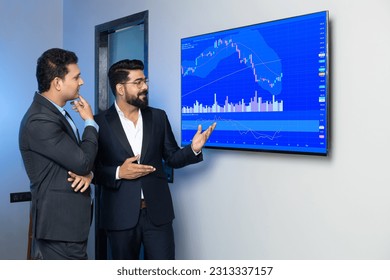Indian businessmen colleagues trading and discussing stocks online in office. Stock brokers looking at indexes graphs on big computer screen.