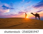 Indian cameleer camel driver bedouin with camel silhouettes in sand dunes of Thar desert on sunset. Caravan in Rajasthan travel tourism background safari adventure. Jaisalmer, Rajasthan, India