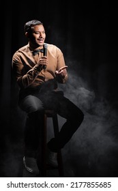 Indian Comedian Sitting And Performing Stand Up Comedy Into Microphone On Black With Smoke