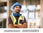 Indian construction site manager standing with folded arms wearing safety vest and helmet, thinking at construction site. Portrait of mixed race manual worker or architect with satisfaction.