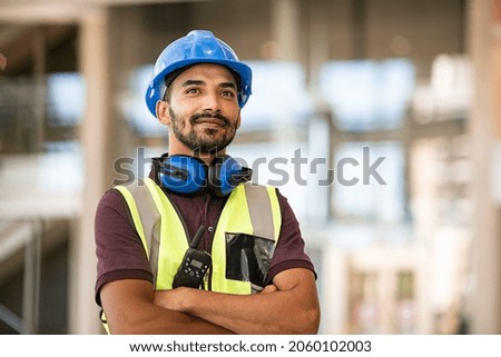 Indian construction site manager standing with folded arms wearing safety vest and helmet, thinking at construction site. Portrait of mixed race manual worker or architect with satisfaction.