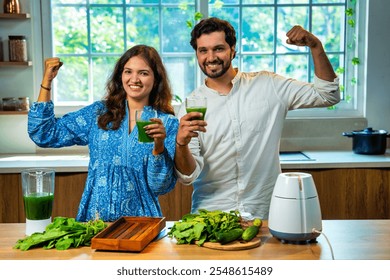 Indian couple preparing fresh green spinach juice together in kitchen, both showing strength gesture promoting health and fitness - Powered by Shutterstock