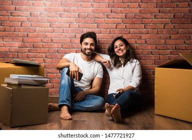 Indian couple relaxing while shifting/unpacking home, sitting between packaging boxes, looking at Photo frame, cel phone