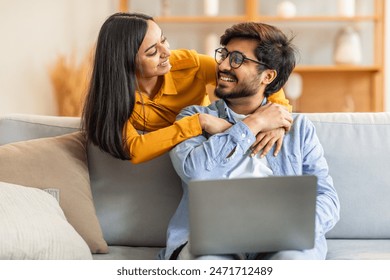 Indian couple is seen in a cozy living room, where they share a heartfelt moment. The man is using a laptop while the woman lovingly embraces him from behind, both smiling warmly. - Powered by Shutterstock