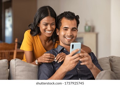 Indian Couple Using Smartphone Together At Home. Man Sitting On Couch Showing Woman A Video. Middle Eastern Girl Embracing From Behind Her Boyfriend While Doing Video Call On Mobile Phone, Copy Space.