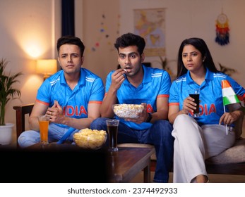 Indian cricket fans sitting on a couch and watching a live cricket match on TV. Friends sitting together in the living room with popcorn, chips, and cold drinks to watch Indian cricket match - cric...