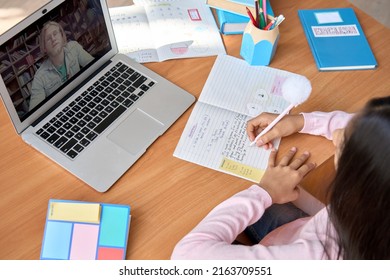 Indian Cute Kid School Girl Having Virtual Online Foreign Language Class With Male Tutor Teacher Sitting At Table At Home. Over Shoulder Top View Of Latin Student Learning Writing In Notebook.