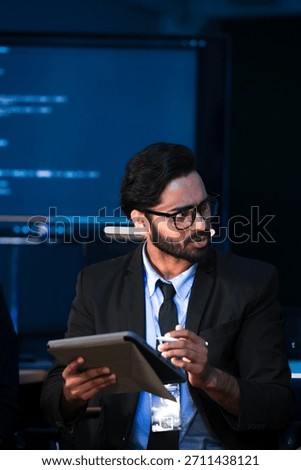 An Indian data scientist explains an AI algorithm on a tablet to a Caucasian software engineer. They are reviewing system data in a cybersecurity command center
