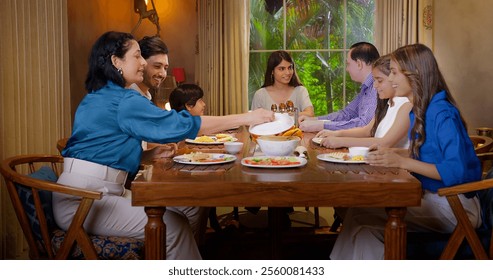 Indian diverse extended happy family group sitting around dining table chair have night dinner together. Smiling young adult children with older parent talk gossip eating fresh salad enjoy spend time - Powered by Shutterstock