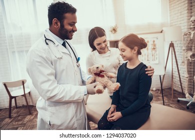 Indian Doctor In White Gown Seeing Patients In Office. Doctor Is Giving Mother And Daughter Teddy Bear.