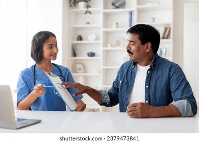 Indian Doctor Woman Showing Clipboard With Medical Survey To Mature Male Patient, Professional Therapist Lady Discussing Therapy Process With Middle Aged Man During Appointment In Office - Powered by Shutterstock