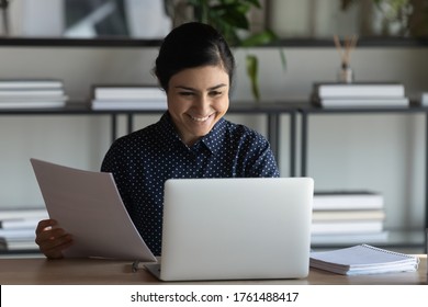 Indian employee do work seated at workplace desk holding bank receipt sheet tax refund notification, paper bill letter, read good news check e-mail on pc, accomplish paperwork feels satisfied concept
