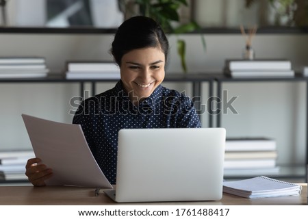 Indian employee do work seated at workplace desk holding bank receipt sheet tax refund notification, paper bill letter, read good news check e-mail on pc, accomplish paperwork feels satisfied concept