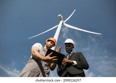 Indian engineer in uniform and helmet using tablet during meeting with african american inspector in stylish suit. Two partners discussing green energy while standing on farm with windmills. - Powered by Shutterstock