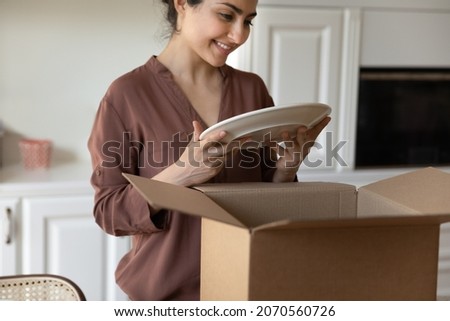 Indian ethnicity woman holds plate while unpack parcel box with delivered, ordered online in e-commerce retail services web stores kitchen quality items. Satisfied client buyer of new crockery concept