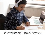 Indian ethnicity woman wearing headphones listens educational course writing down necessary useful information gain knowledge online use internet lesson, interpreter do translation sit at desk indoors