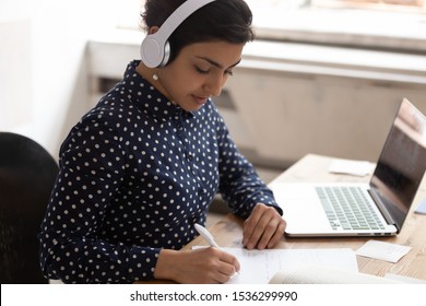 Indian ethnicity woman wearing headphones listens educational course writing down necessary useful information gain knowledge online use internet lesson, interpreter do translation sit at desk indoors