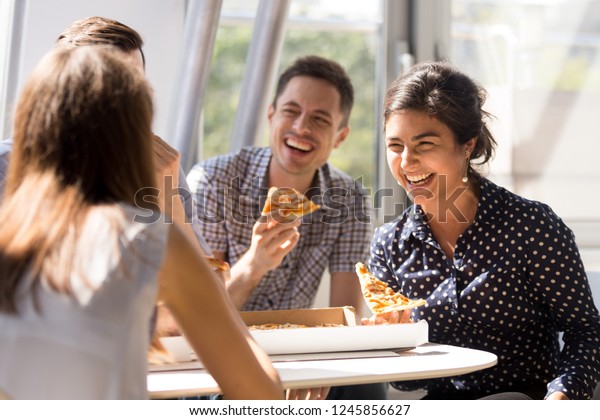 Indian excited woman laughing at funny joke,\
eating pizza with diverse colleagues in office, happy multi-ethnic\
employees having fun together during lunch, enjoying good\
conversation, emotions